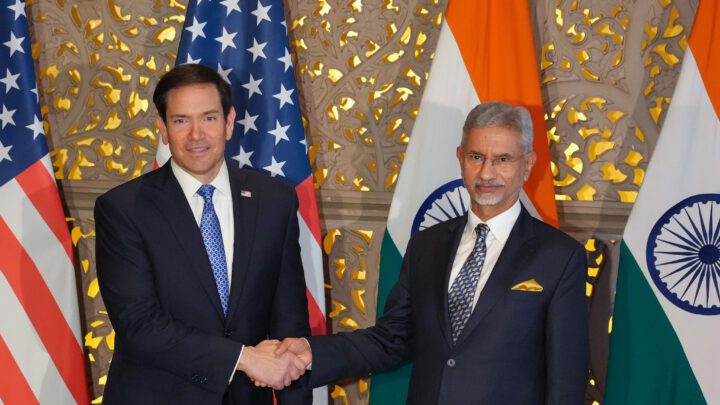 Marco Rubio and S Jaishankar, both in dark suits, shake hands in front of US and Indian flags and a gold latticed wall.