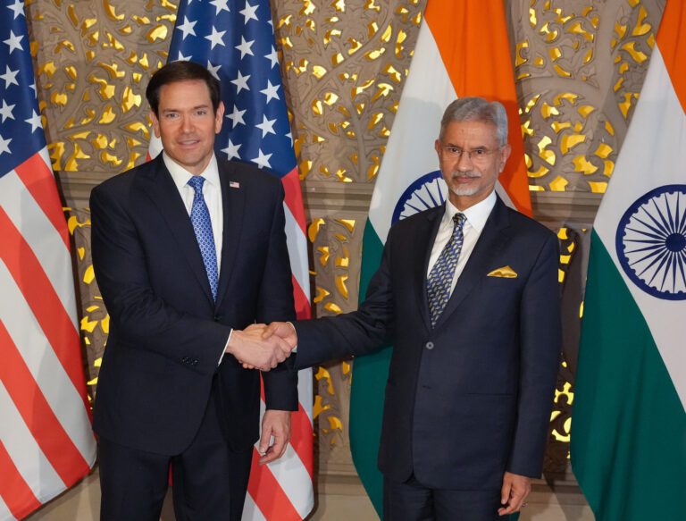 Marco Rubio and S Jaishankar, both in dark suits, shake hands in front of US and Indian flags and a gold latticed wall.