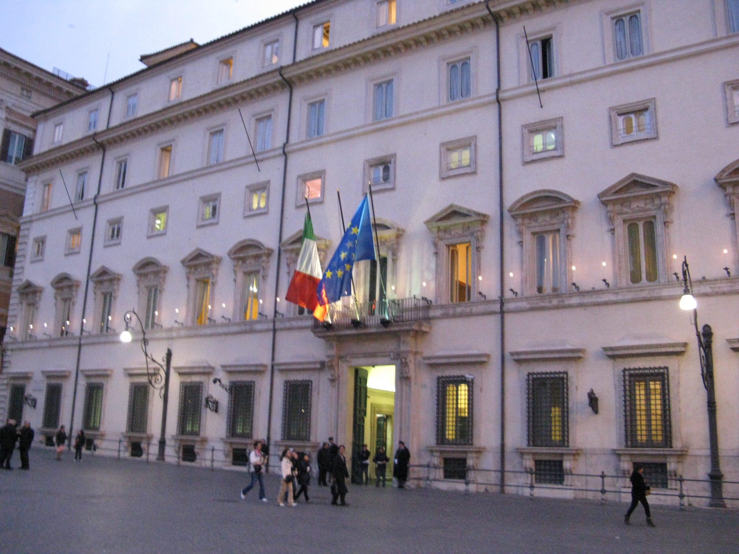 The pale stone facade of Palazzo Chigi in Rome at dusk, with Italian and European Union flags over the main entrance and a few people walking across the square.
