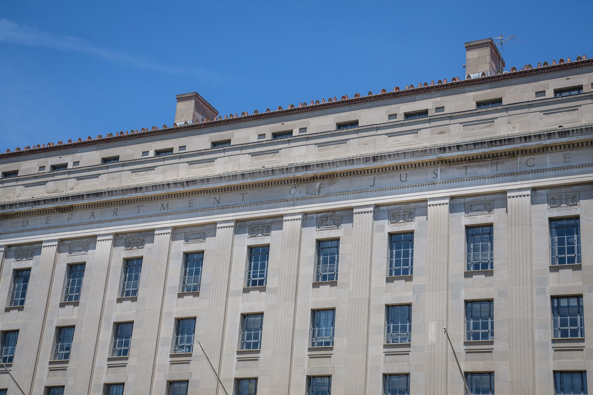 The upper floors of a pale limestone building with rows of windows and the words Department of Justice carved across the facade, under a blue sky.