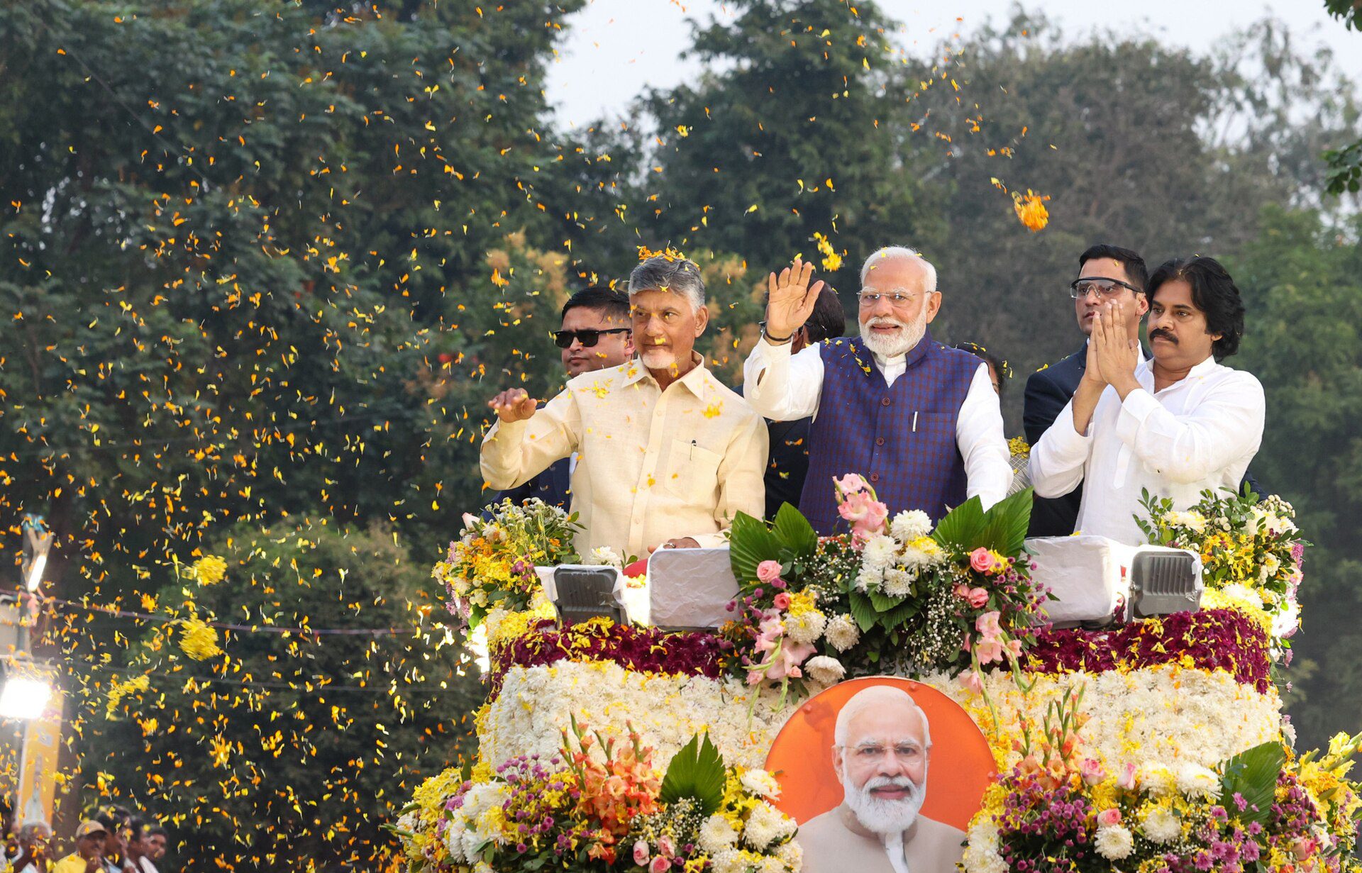 Chandrababu Naidu, Narendra Modi and Pawan Kalyan wave and greet the crowd from a flower-decked open vehicle as petals fall around them.