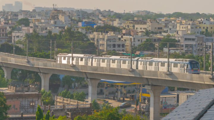 A white three-coach metro train running on an elevated concrete viaduct above a street, with residential buildings behind it.