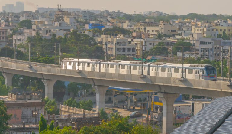 A white three-coach metro train running on an elevated concrete viaduct above a street, with residential buildings behind it.