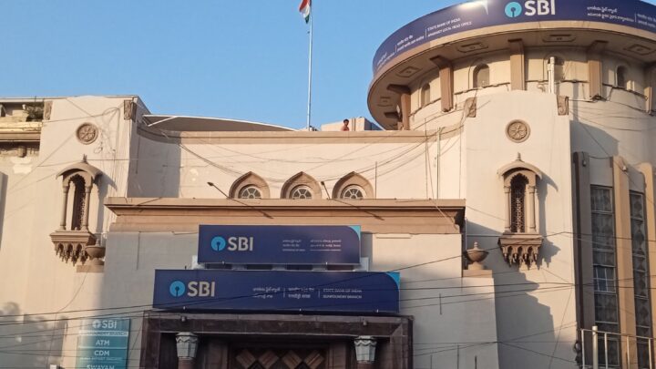 A cream-coloured stone building with blue SBI signboards above a carved wooden doorway, a rounded tower carrying another SBI sign and the national flag on the roof.