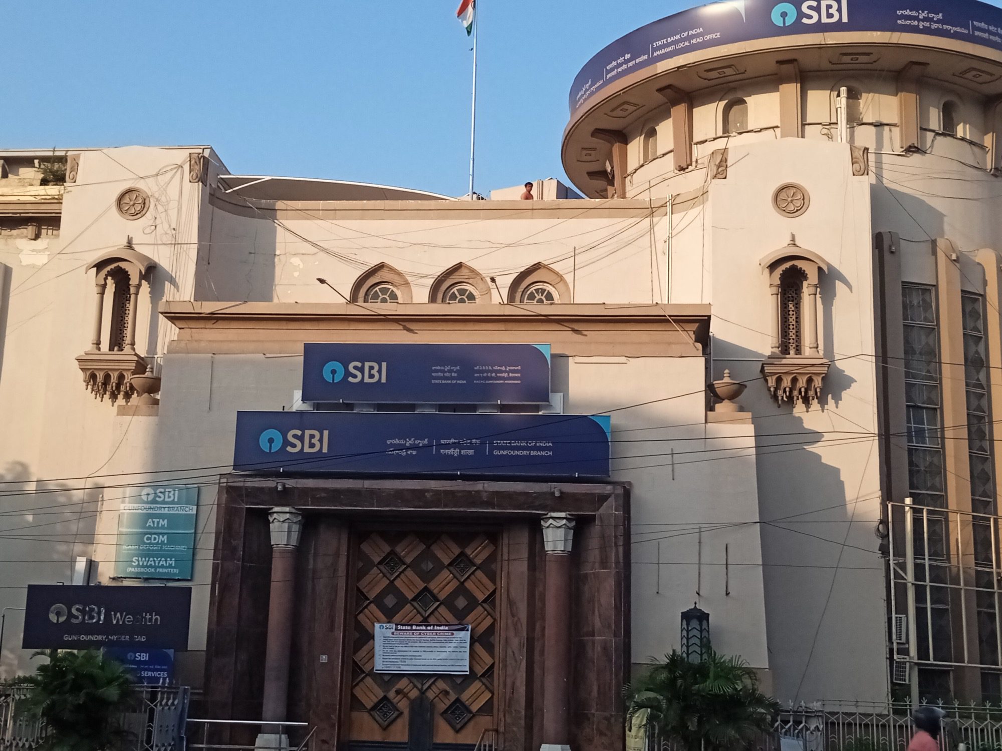 A cream-coloured stone building with blue SBI signboards above a carved wooden doorway, a rounded tower carrying another SBI sign and the national flag on the roof.