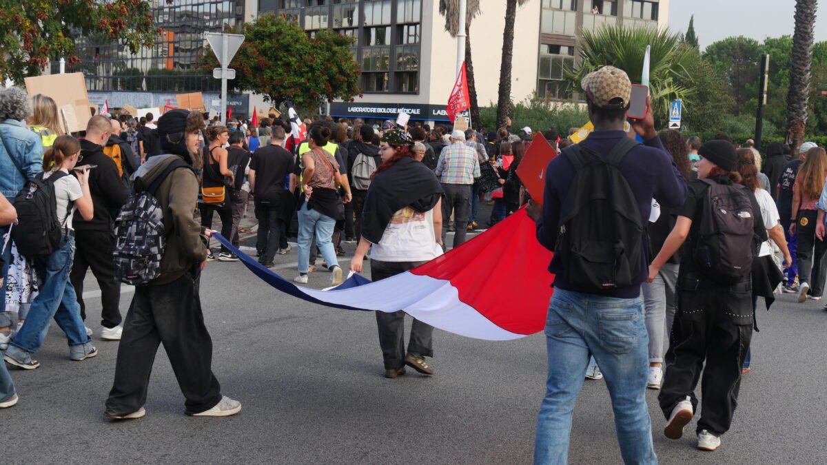 Young people with backpacks march down a city street, several of them carrying a long French tricolour flag, with placards and office buildings in the background.