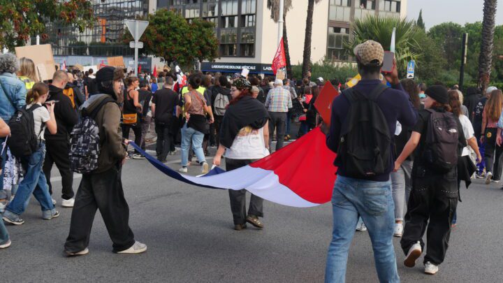 Young people with backpacks march down a city street, several of them carrying a long French tricolour flag, with placards and office buildings in the background.