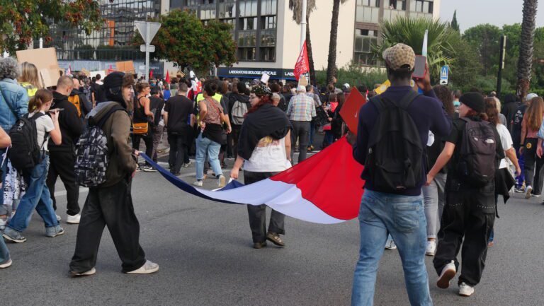 Young people with backpacks march down a city street, several of them carrying a long French tricolour flag, with placards and office buildings in the background.