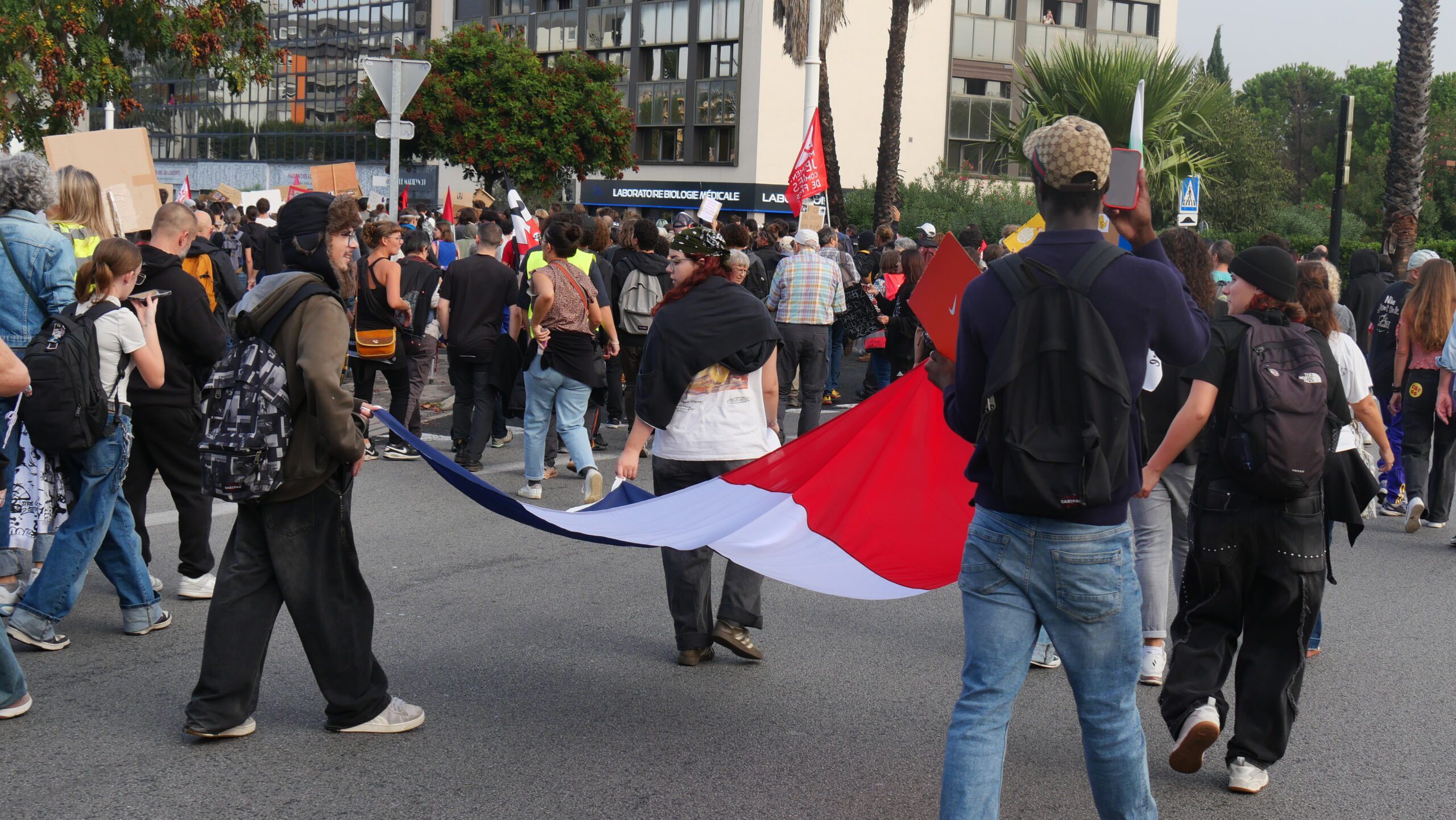 Young people with backpacks march down a city street, several of them carrying a long French tricolour flag, with placards and office buildings in the background.