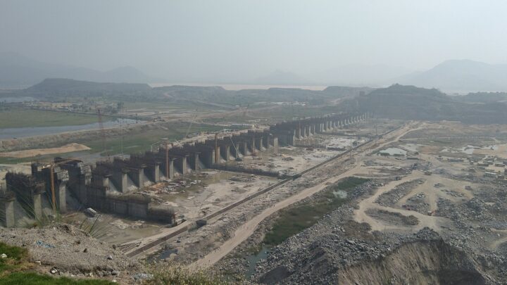Concrete spillway piers of the Polavaram dam under construction, with cranes, hills and a river in the background.