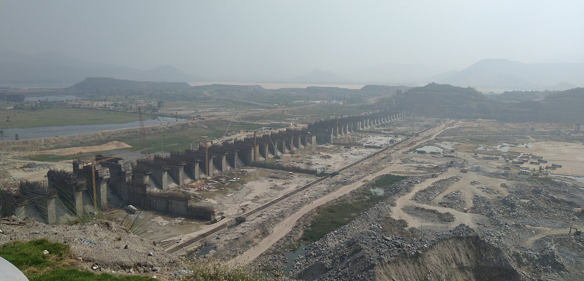 Concrete spillway piers of the Polavaram dam under construction, with cranes, hills and a river in the background.