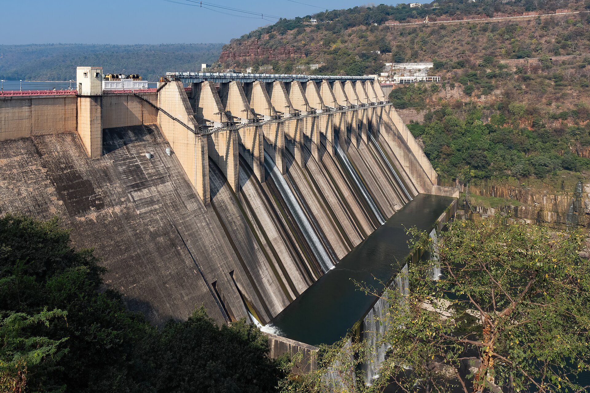 Concrete spillway gates and buttresses of Srisailam dam with water streaming down two channels, hills behind