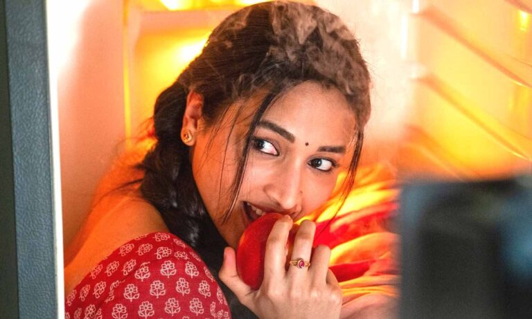 Srinidhi Shetty as Swarna in Aadarsha Kutumbam, biting an apple at an open fridge