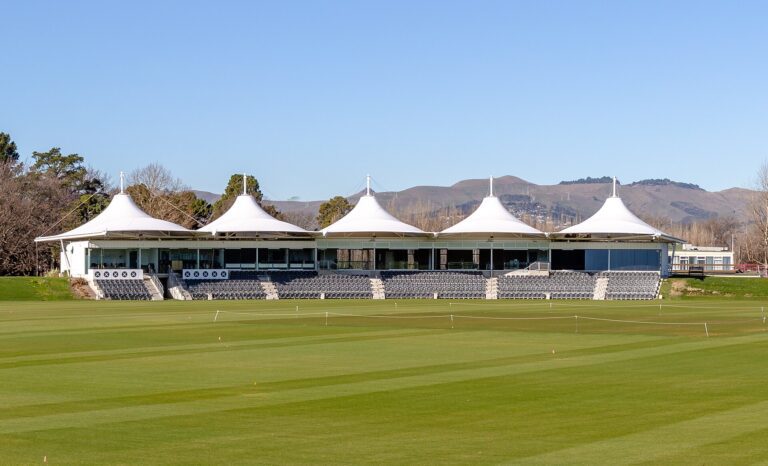 White tent-roofed pavilion and grey stands behind a green cricket outfield at Hagley Oval, with hills in the background.