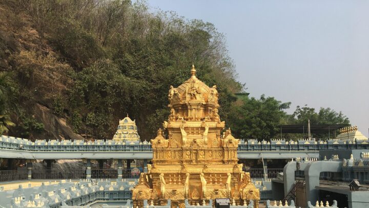 Gold-plated tower of the Kanaka Durga temple with temple walls and a wooded hillside behind it