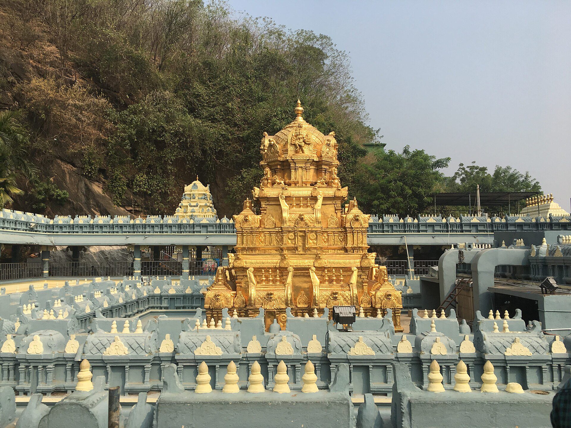 Gold-plated tower of the Kanaka Durga temple with temple walls and a wooded hillside behind it