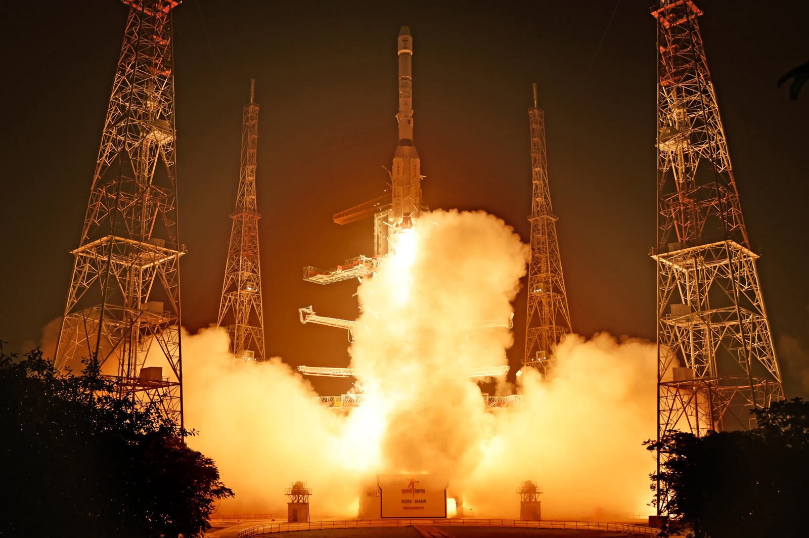 GSLV-F18 rocket rising from the launch pad at night amid bright exhaust clouds, flanked by lightning towers