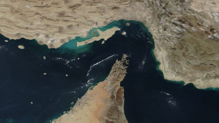 Satellite view of the Strait of Hormuz with the Musandam Peninsula jutting into dark blue water between Iran and the Arabian coast.