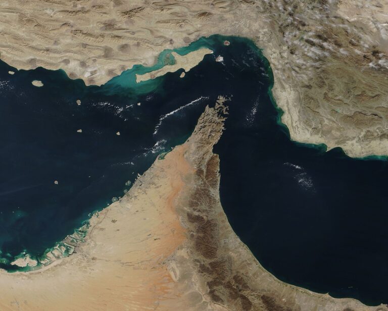Satellite view of the Strait of Hormuz with the Musandam Peninsula jutting into dark blue water between Iran and the Arabian coast.