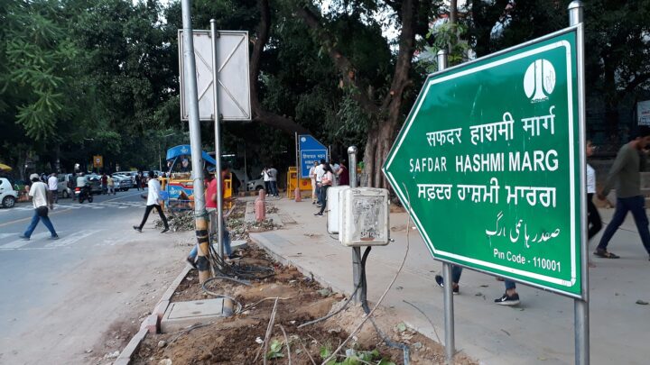 A green road sign reading Safdar Hashmi Marg in Hindi, English, Punjabi and Urdu beside a tree-lined Delhi road, with pedestrians and parked vehicles in the background.