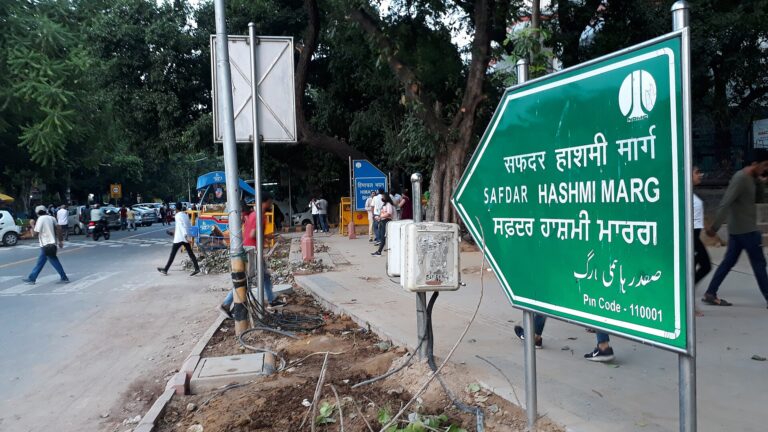 A green road sign reading Safdar Hashmi Marg in Hindi, English, Punjabi and Urdu beside a tree-lined Delhi road, with pedestrians and parked vehicles in the background.
