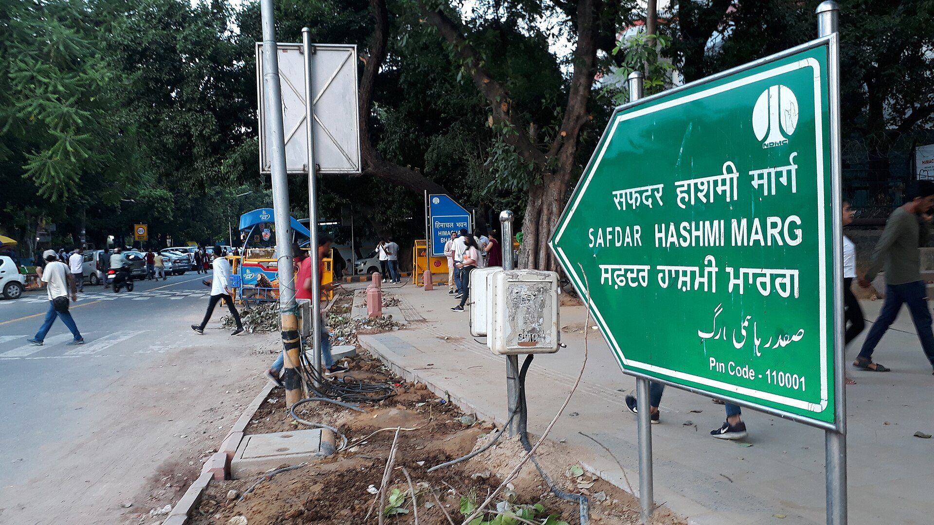 A green road sign reading Safdar Hashmi Marg in Hindi, English, Punjabi and Urdu beside a tree-lined Delhi road, with pedestrians and parked vehicles in the background.