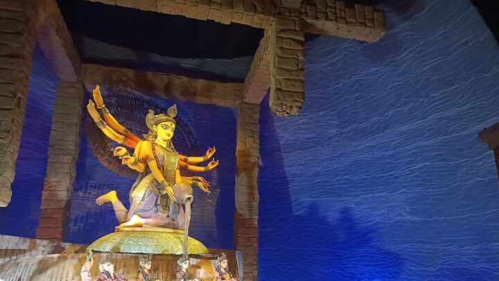 A Durga idol on a lotus base, pouring water from a pot, inside a pandal with brick-style pillars and a blue wave backdrop.