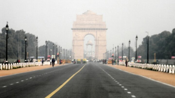 India Gate at the end of a wide road in Delhi, partly veiled in grey haze, with a few people and auto-rickshaws in the distance