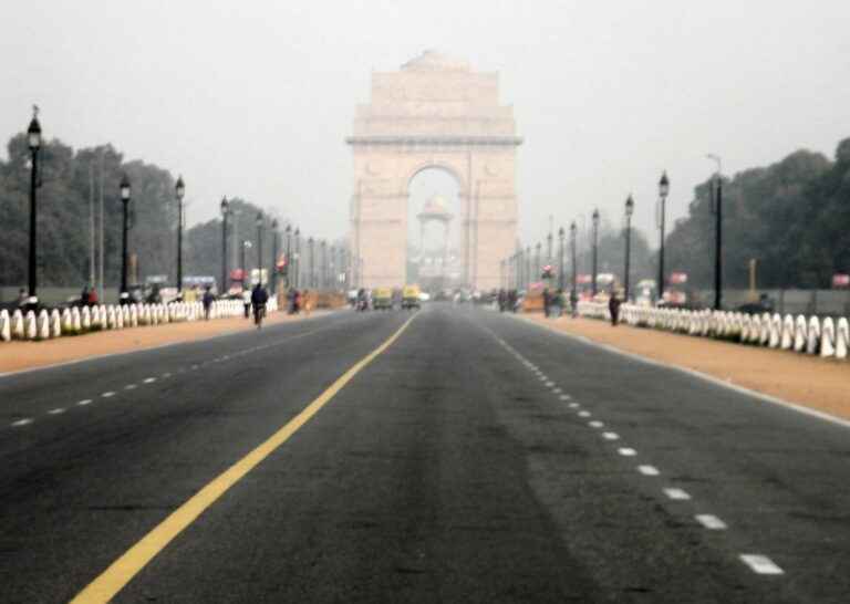 India Gate at the end of a wide road in Delhi, partly veiled in grey haze, with a few people and auto-rickshaws in the distance