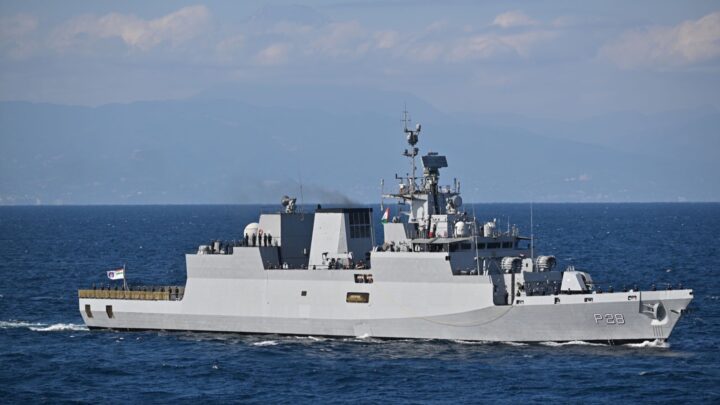 Indian Navy corvette INS Kamorta, hull number P28, sailing on a calm blue sea with crew standing along the deck and hazy mountains behind.