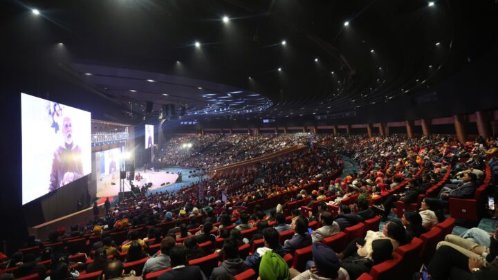 A packed auditorium in New Delhi with rows of red seats facing a stage, and a large screen showing a speaker at the podium.