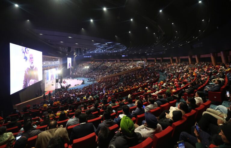 A packed auditorium in New Delhi with rows of red seats facing a stage, and a large screen showing a speaker at the podium.