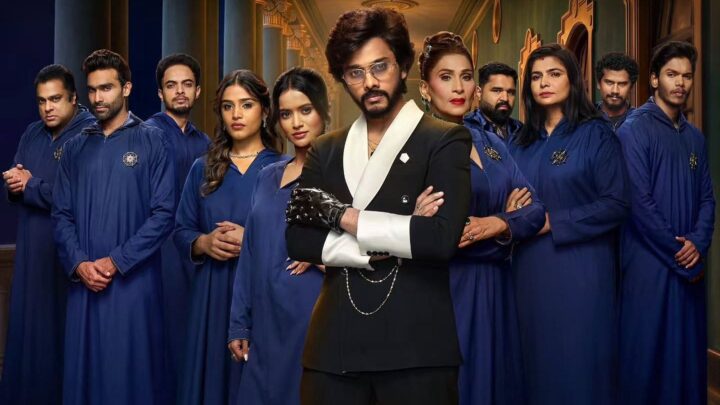 Host Teja Sajja in a black-and-white suit, standing in front of contestants dressed in matching blue robes.