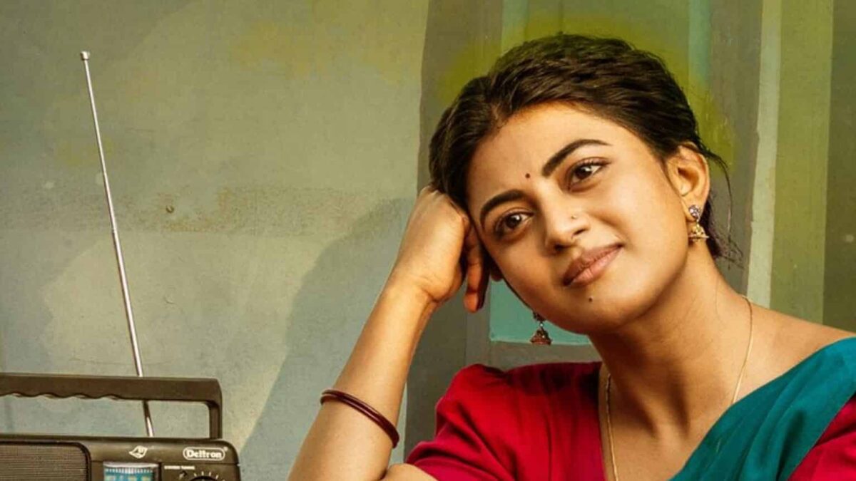 Anandhi, in a red blouse and green sari, rests her head on her hand beside an old transistor radio.