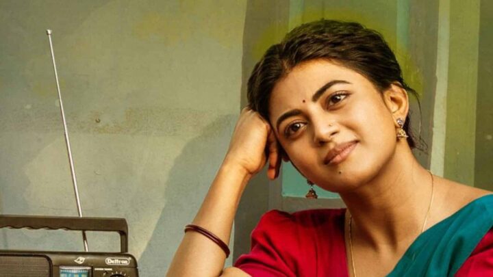 Anandhi, in a red blouse and green sari, rests her head on her hand beside an old transistor radio.