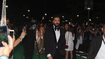Actor Riteish Deshmukh, in a black tuxedo and bow tie, walks along a green carpet at night as fans take photos on their phones.