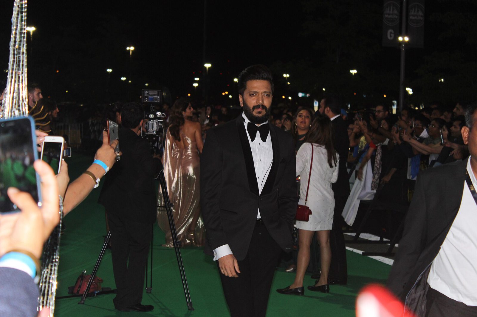 Actor Riteish Deshmukh, in a black tuxedo and bow tie, walks along a green carpet at night as fans take photos on their phones.