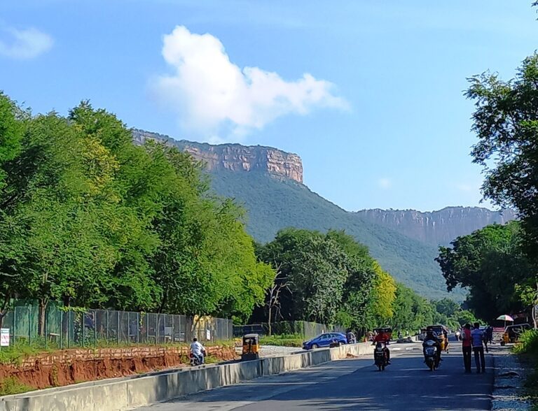 Forested Tirumala hills with a rocky escarpment under a blue sky, seen from a tree-lined road with a few motorcycles and autorickshaws.