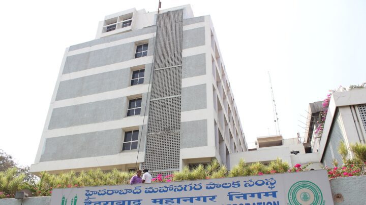 Multi-storey Greater Hyderabad Municipal Corporation head office with its blue trilingual signboard and flowering plants along the front.