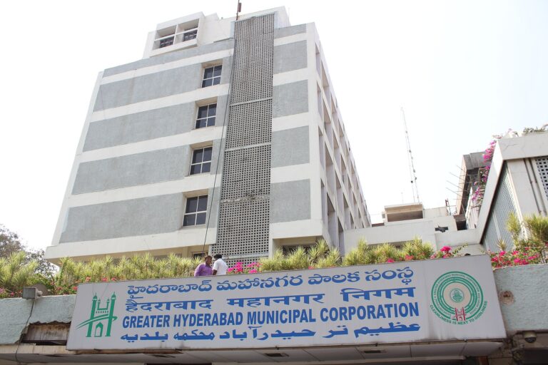 Multi-storey Greater Hyderabad Municipal Corporation head office with its blue trilingual signboard and flowering plants along the front.