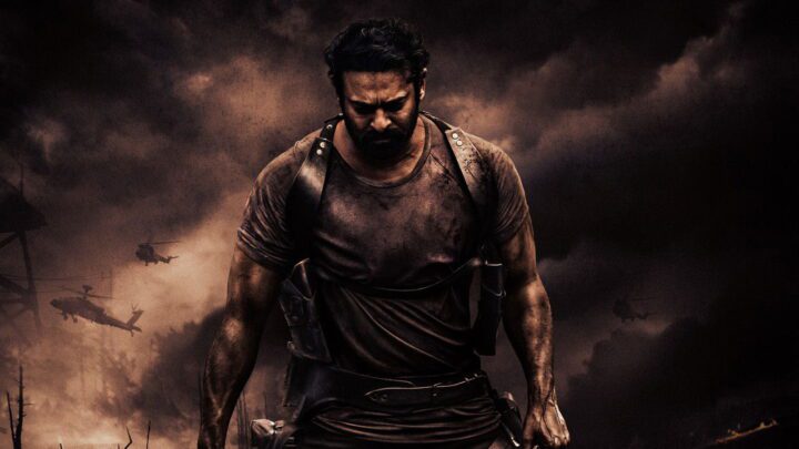 Prabhas as Salaar, bearded and in a mud-stained vest with leather holsters, walking under a dark smoky sky with helicopters behind him.