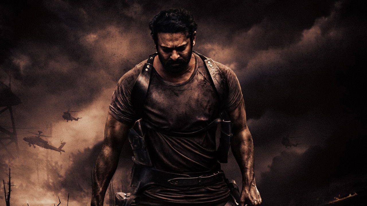 Prabhas as Salaar, bearded and in a mud-stained vest with leather holsters, walking under a dark smoky sky with helicopters behind him.