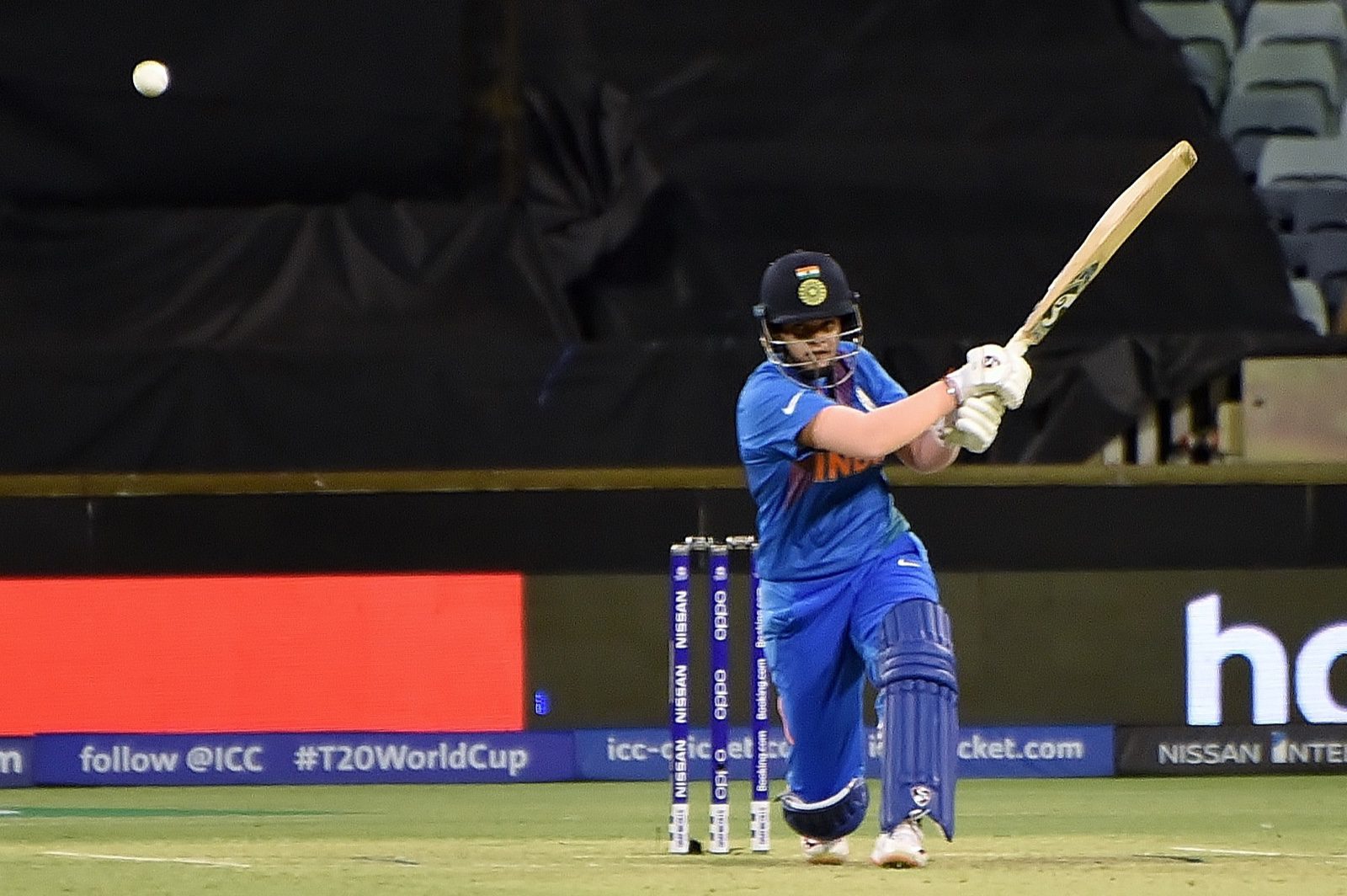 Indian batter Shafali Verma, in a blue India kit and helmet, plays a shot in front of the stumps at a floodlit cricket ground.