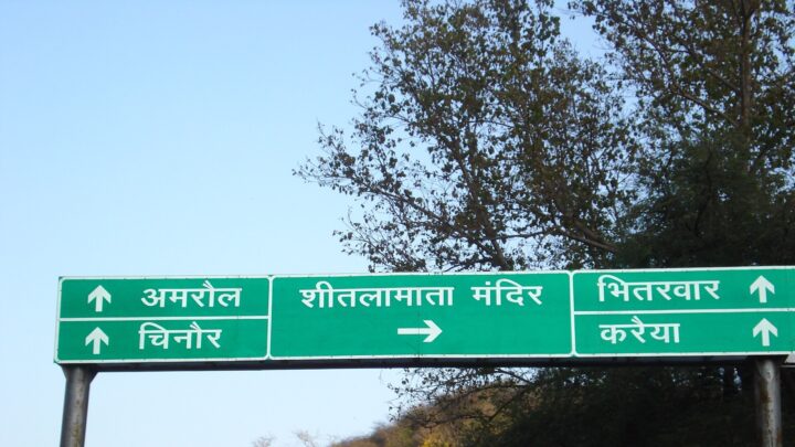 A green overhead road sign in Hindi points right to the Sheetla Mata temple and straight ahead to Amrol, Chinor, Bhitarwar and Karaiya, with a wooded hill behind it.