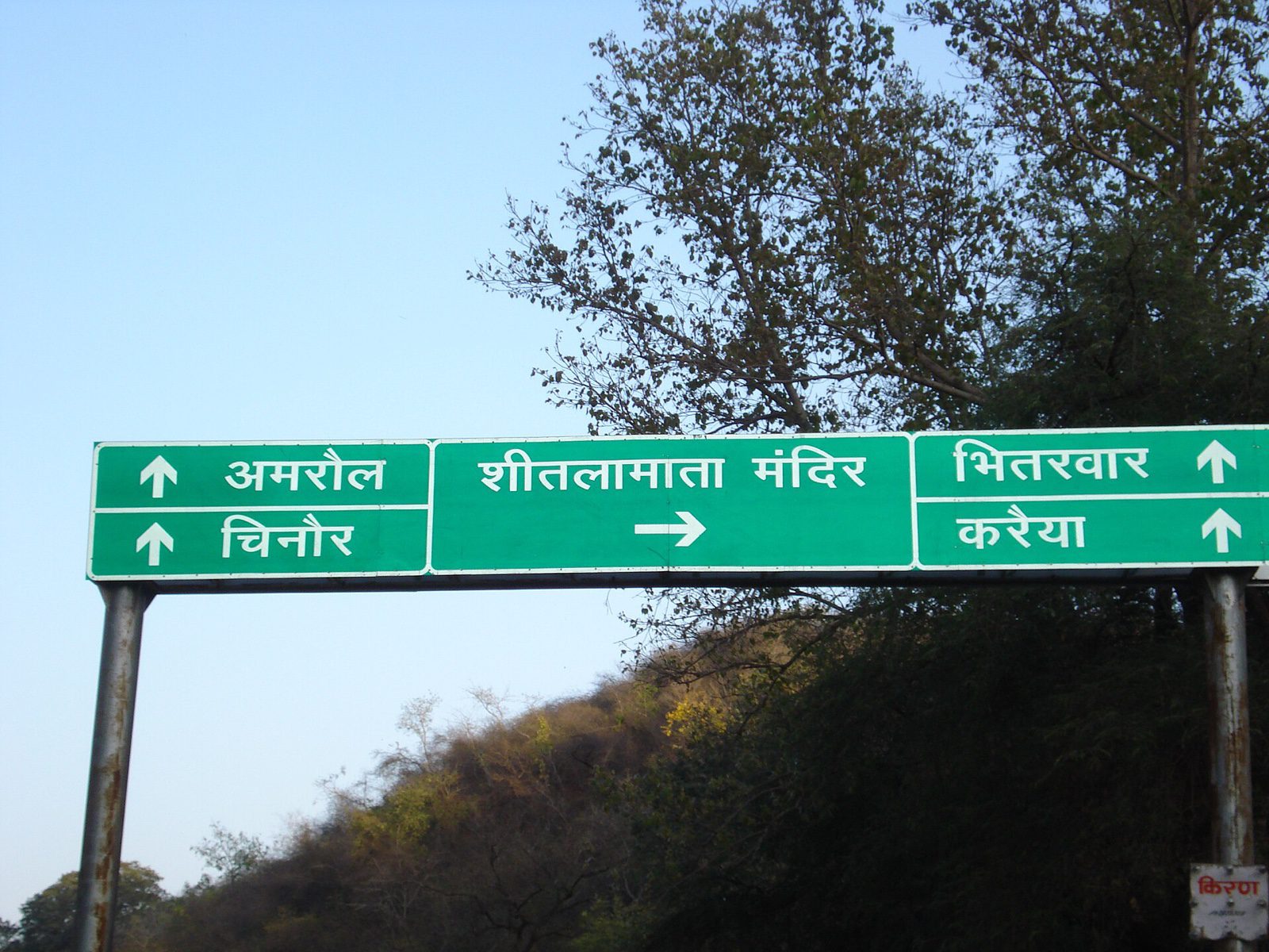 A green overhead road sign in Hindi points right to the Sheetla Mata temple and straight ahead to Amrol, Chinor, Bhitarwar and Karaiya, with a wooded hill behind it.