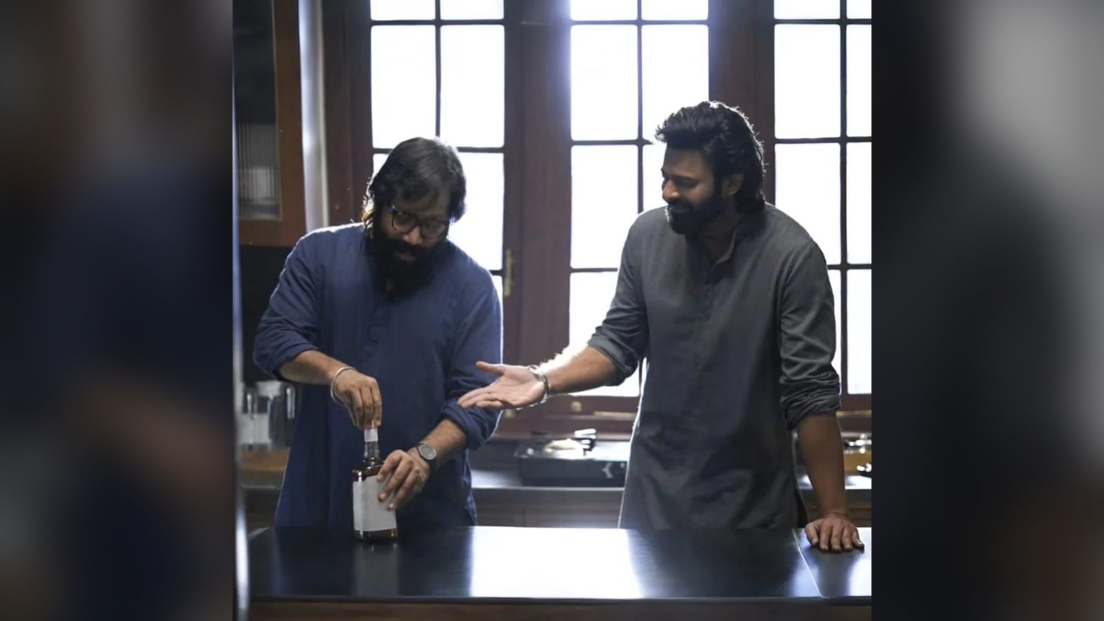 Two men in a kitchen-like room with tall windows: one in a navy shirt and glasses opens a bottle with a plain label, while the other, in a grey kurta, smiles and gestures towards him.