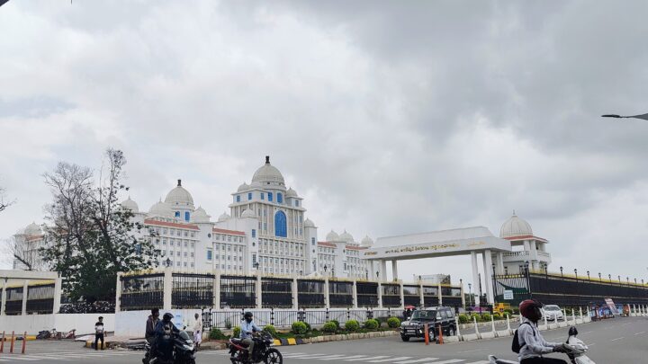 White domed Telangana Secretariat building behind a black iron fence with motorcyclists on the road in front, under a cloudy sky.