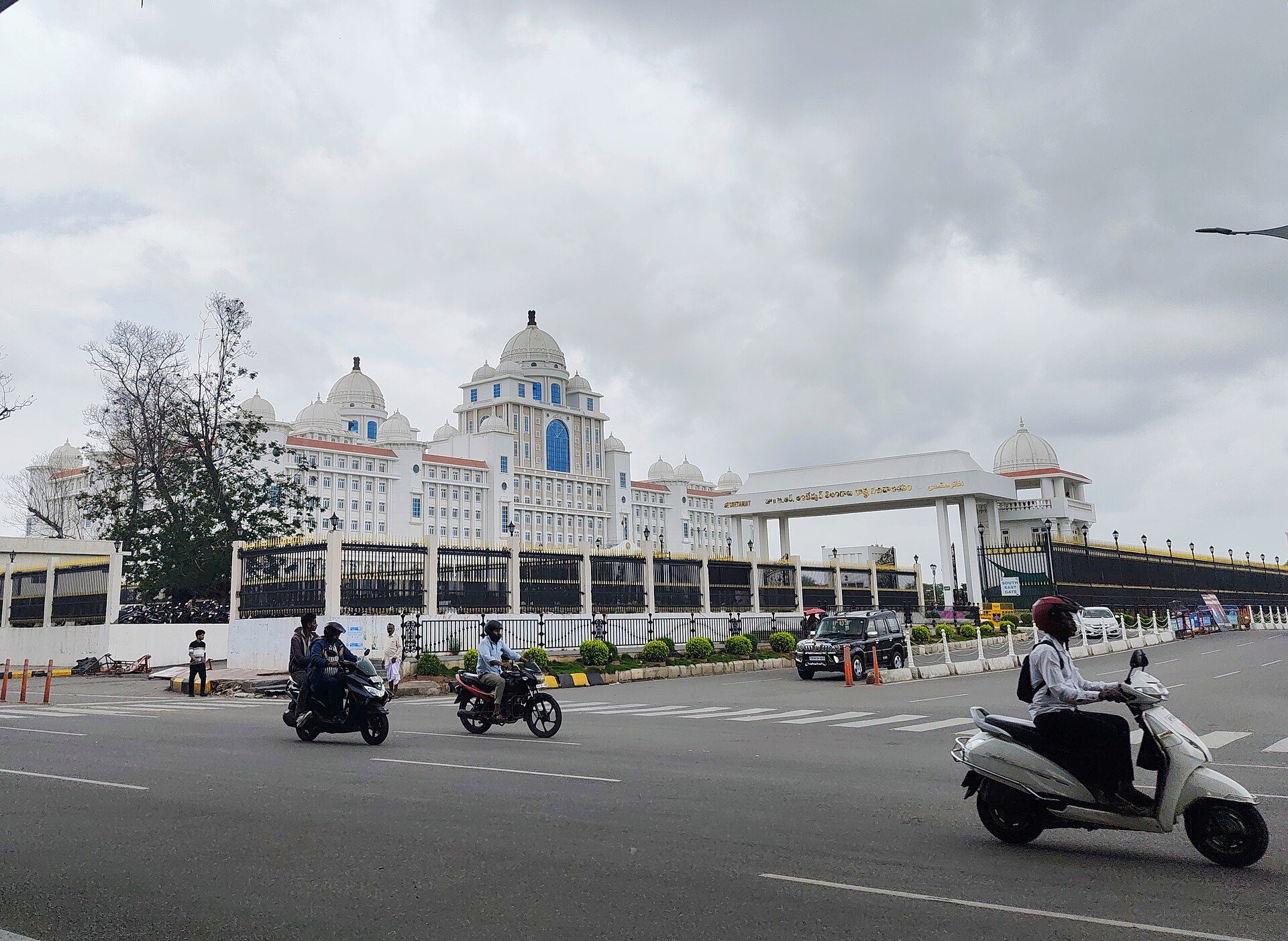 White domed Telangana Secretariat building behind a black iron fence with motorcyclists on the road in front, under a cloudy sky.