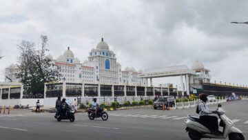White domed Telangana Secretariat building behind a black iron fence, with motorcyclists on the road in front, under a cloudy sky.