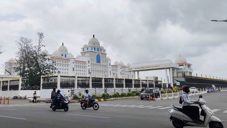 White domed Telangana Secretariat building behind a black iron fence, with motorcyclists on the road in front, under a cloudy sky.